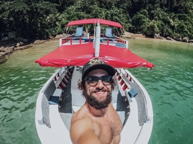 Um homem barbudo, de óculos escuros e boné, sorri em uma selfie em um barco com cobertura vermelha sobre águas calmas e turquesas.