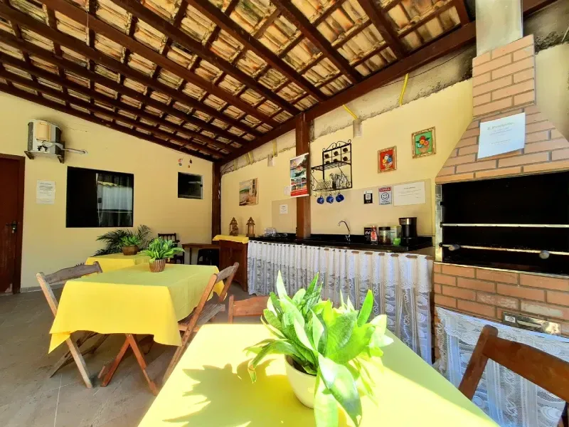 Um pátio externo com mesas cobertas com toalhas amarelas, uma churrasqueira de tijolos e um teto com vigas de madeira.
