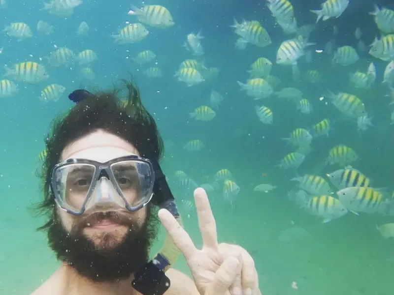 Uma pessoa barbuda usando máscara de mergulho posa debaixo d'água, fazendo o sinal de paz, cercada por um cardume de peixes listrados.
