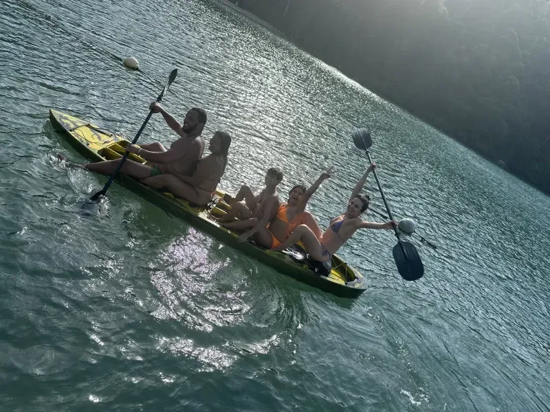 Cinco pessoas em uma prancha de stand-up paddle amarela e comprida em um lago ensolarado, com duas pessoas segurando remos e acenando.