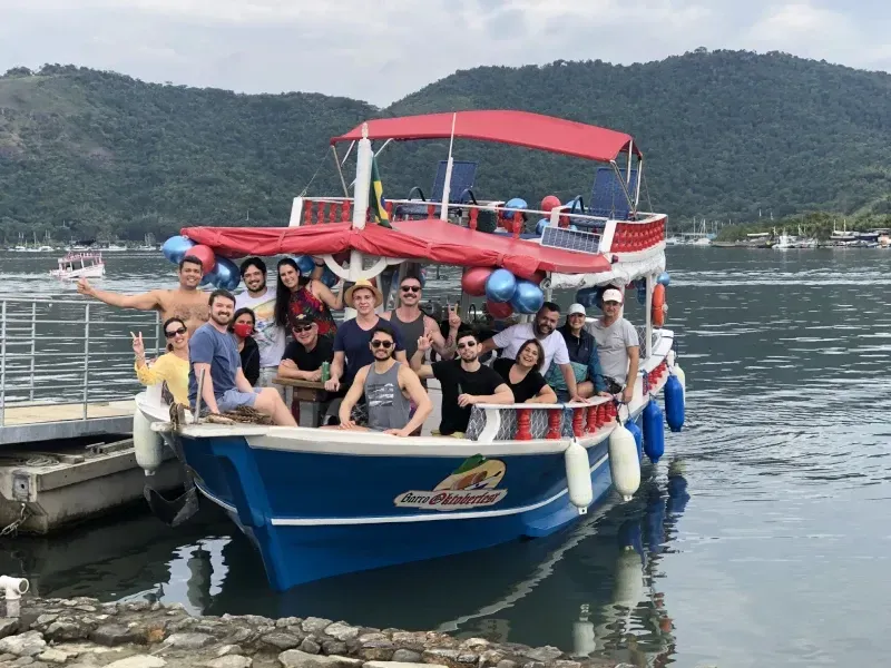 Um grupo de pessoas posa para foto em um barco turístico azul e branco decorado com balões azuis e vermelhos, atracado em um cais em um dia ensolarado.