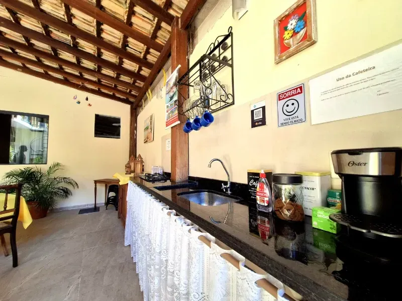 Uma bancada de cozinha com pia, cafeteira e canecas penduradas em uma área de estar externa aberta, com teto de telhas.