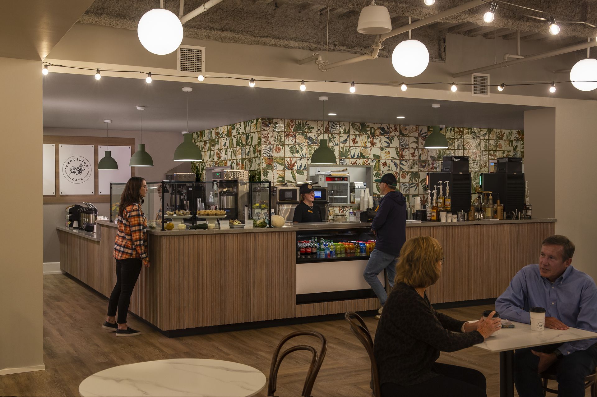 Coffee shop interior with customers and staff at counter. Overhead lights and wood-tone accents.