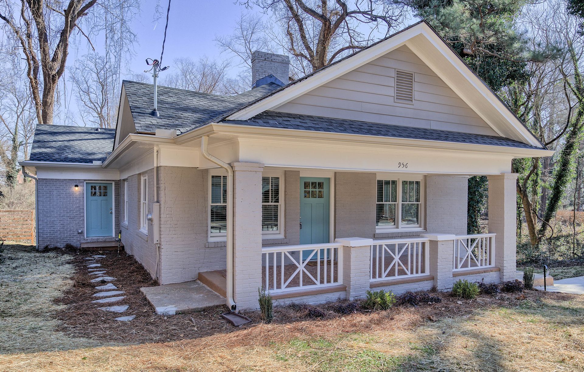 A light-colored bungalow with a porch and teal doors, surrounded by trees.