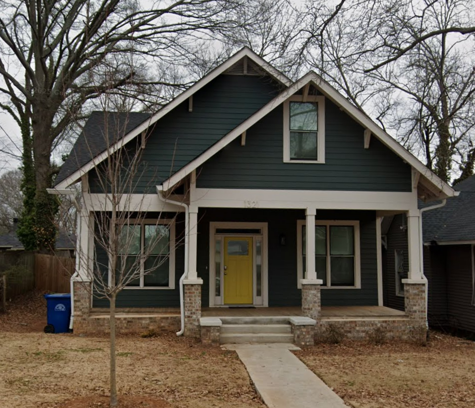 Cottage-style home with dark teal siding, yellow front door, and a stone porch under a cloudy sky.