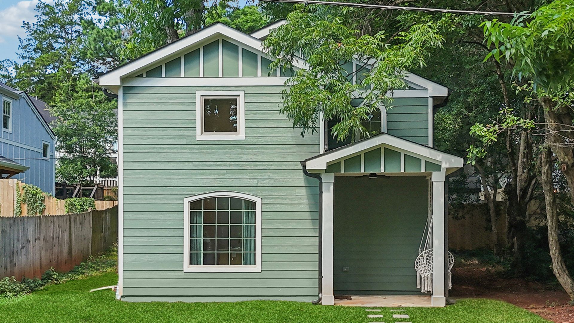Green two-story house with white trim. Porch on the right, trees surround it, and a wooden fence on the left.