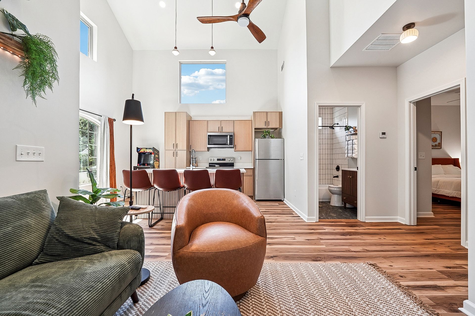 Living room with kitchen in the background. High ceilings, hardwood floors, modern furniture, and natural light.