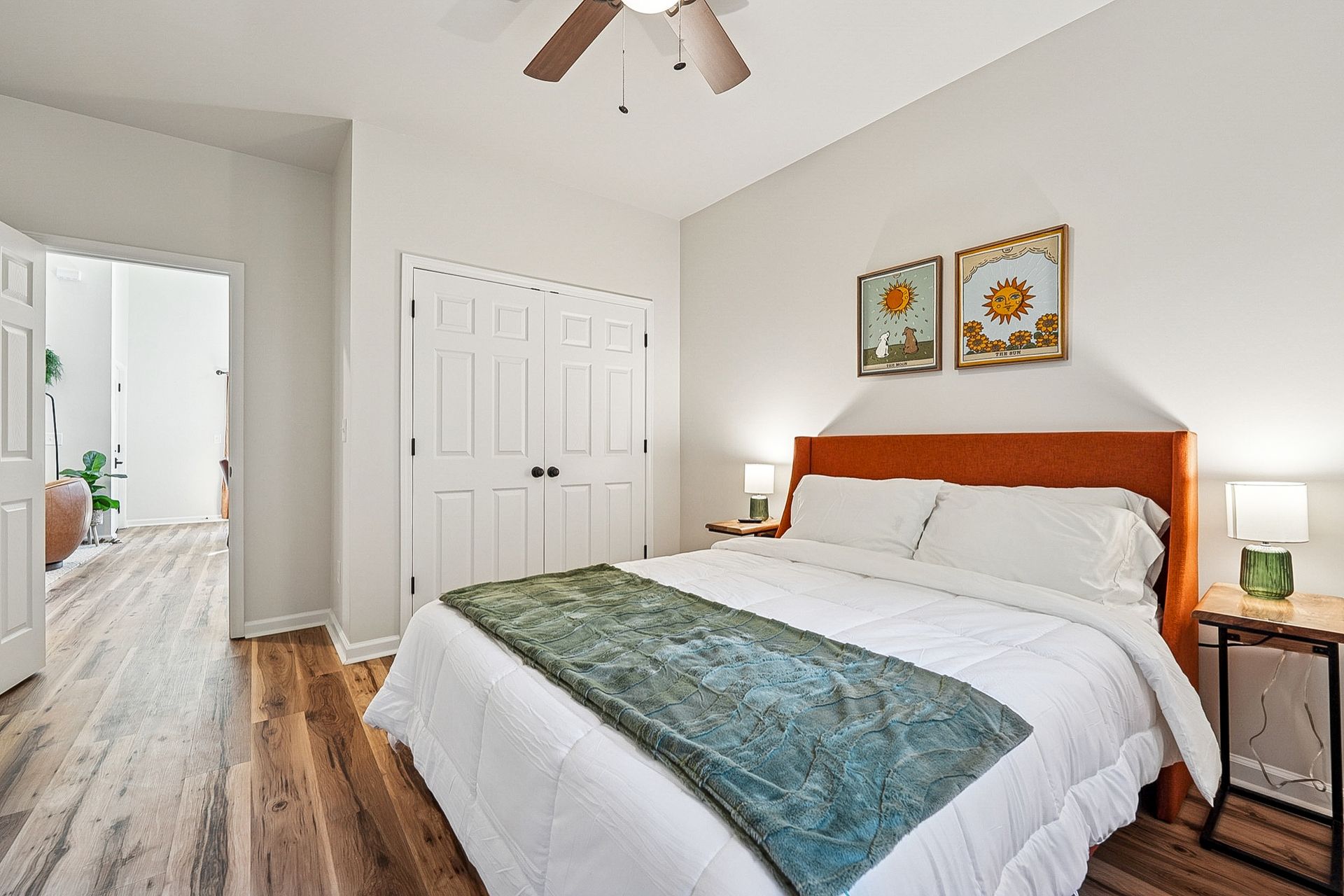 Bedroom with orange headboard, white bedding, green throw, wooden floors, and two sunflower art prints.