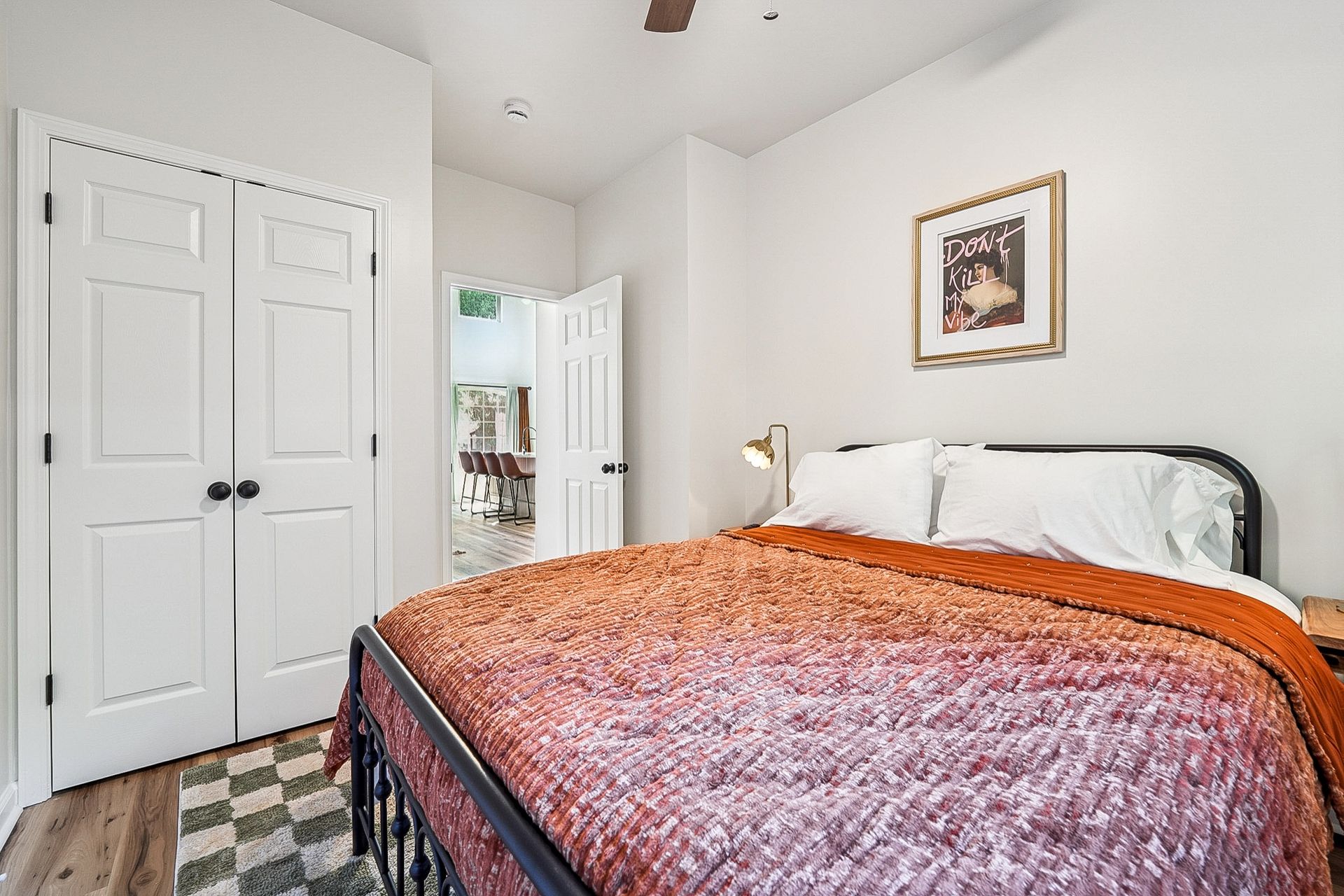 Bedroom with bed, closet doors, open door to another room, artwork, white walls, and wood flooring.