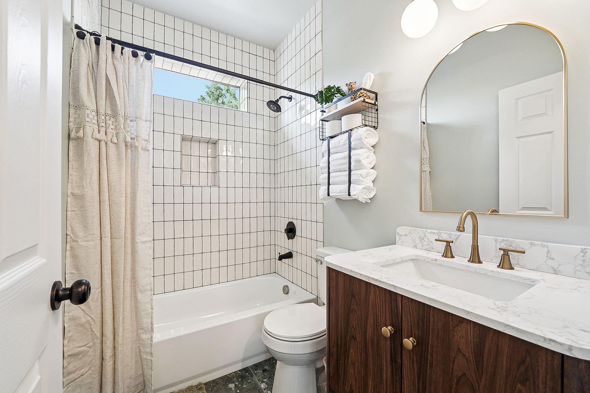 Bathroom with white and black tile, gold accents, dark wood vanity, and arched mirror.