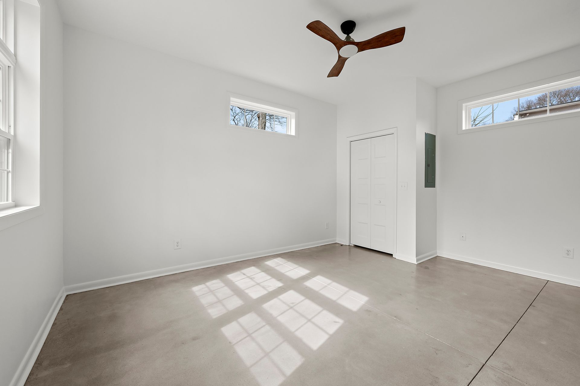 Empty room with white walls, gray floor, ceiling fan, and windows casting shadows.