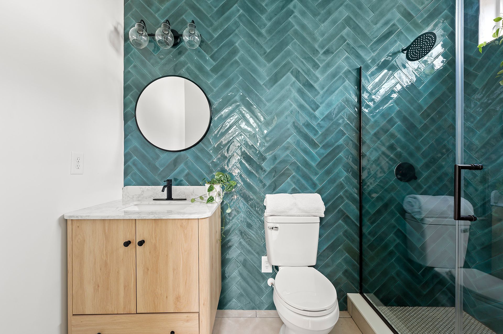 Bathroom with wood vanity, teal herringbone tile wall, round mirror, and glass shower.