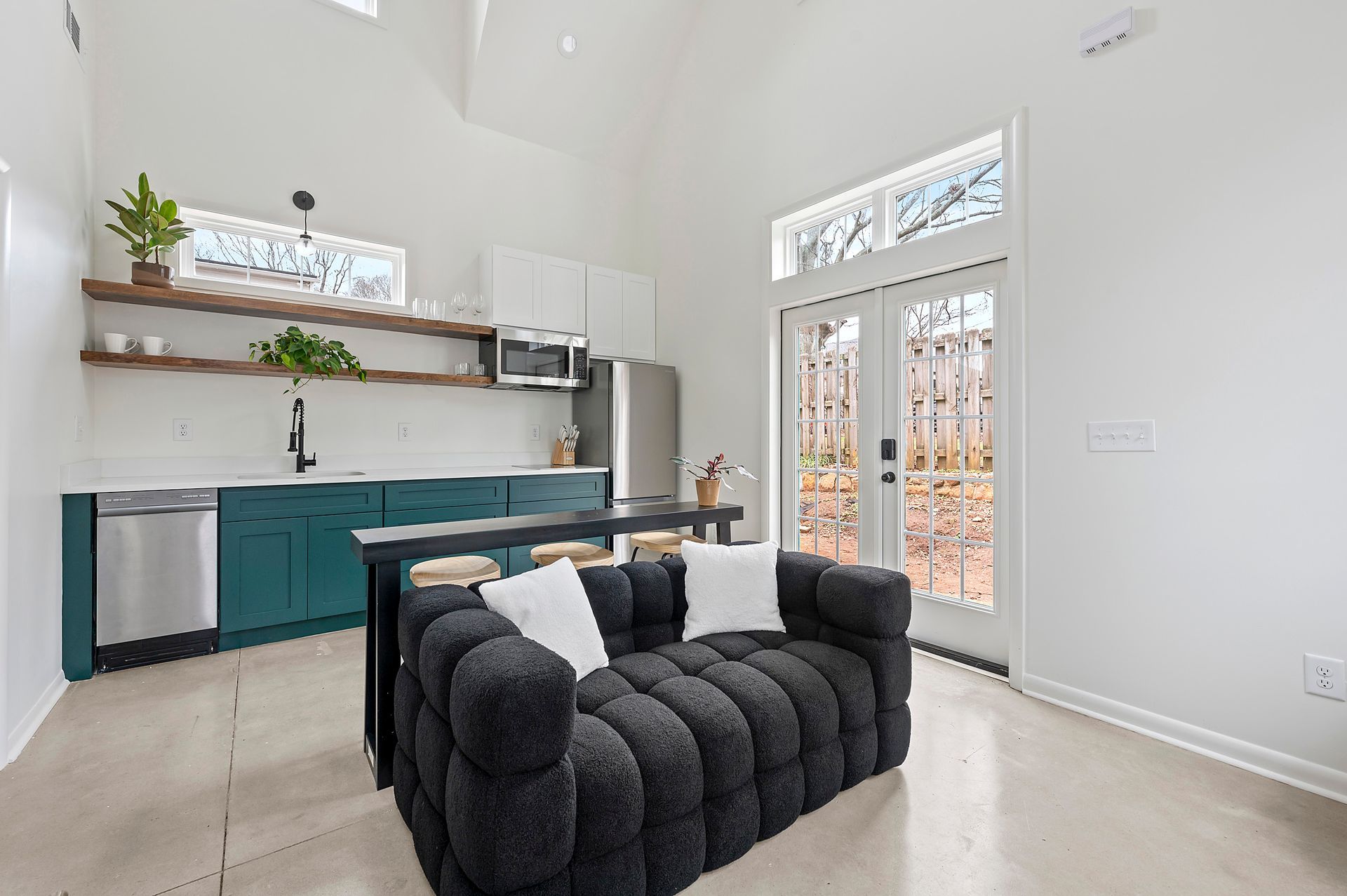 Modern studio apartment with teal cabinets, a black sofa, and a door to a wooded area.