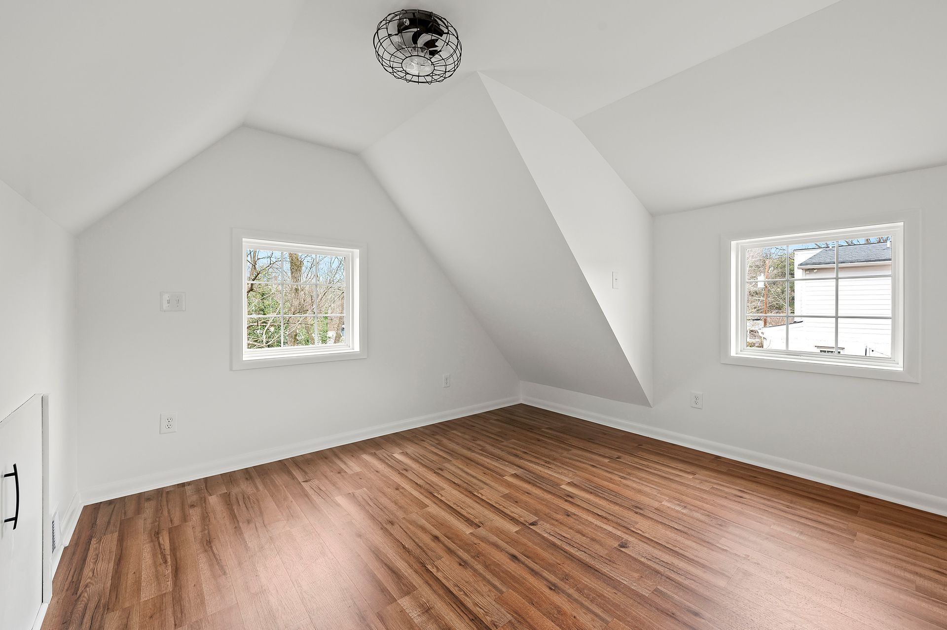 Empty attic room with white walls, wood floor, two windows, and a light fixture.