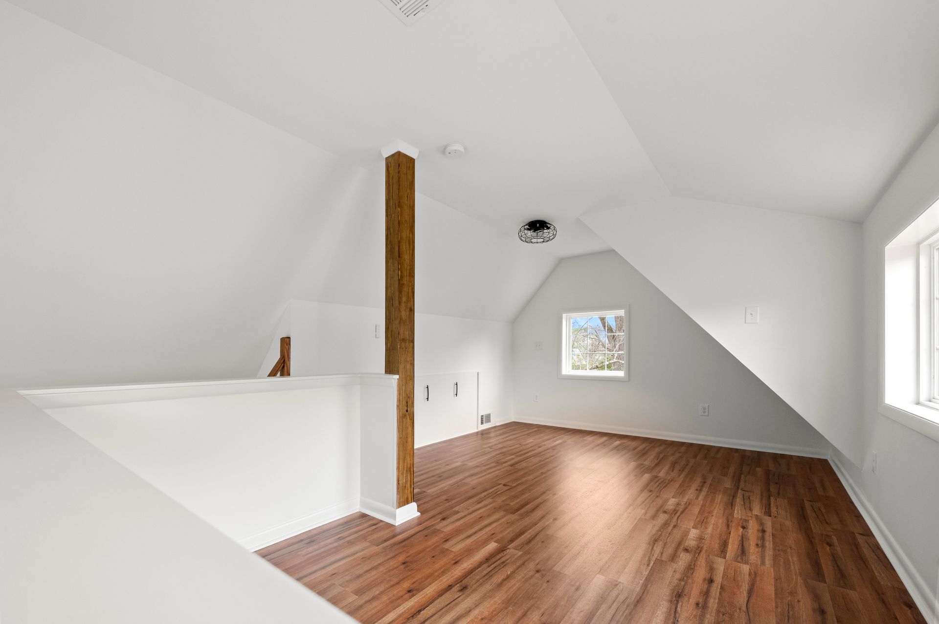Bright, empty attic room with hardwood floor, angled ceiling, small window, and wood beam support.