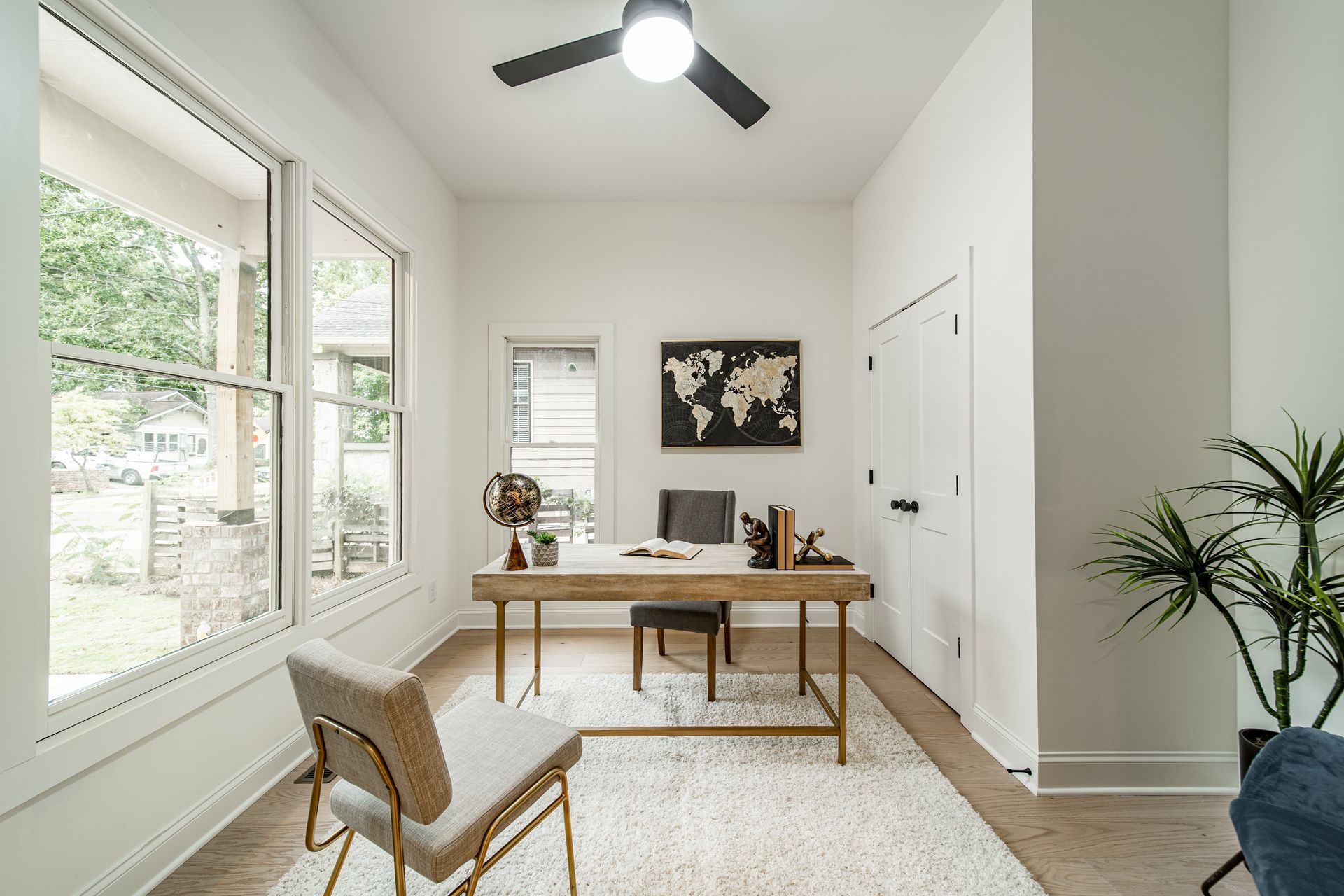 Home office with desk, chair, rug, map, and large windows. White walls and wooden floor.