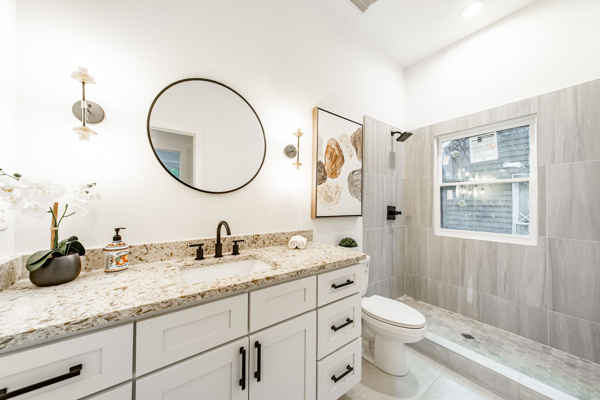 Modern bathroom with white cabinets, speckled countertop, round mirror, and tiled shower.