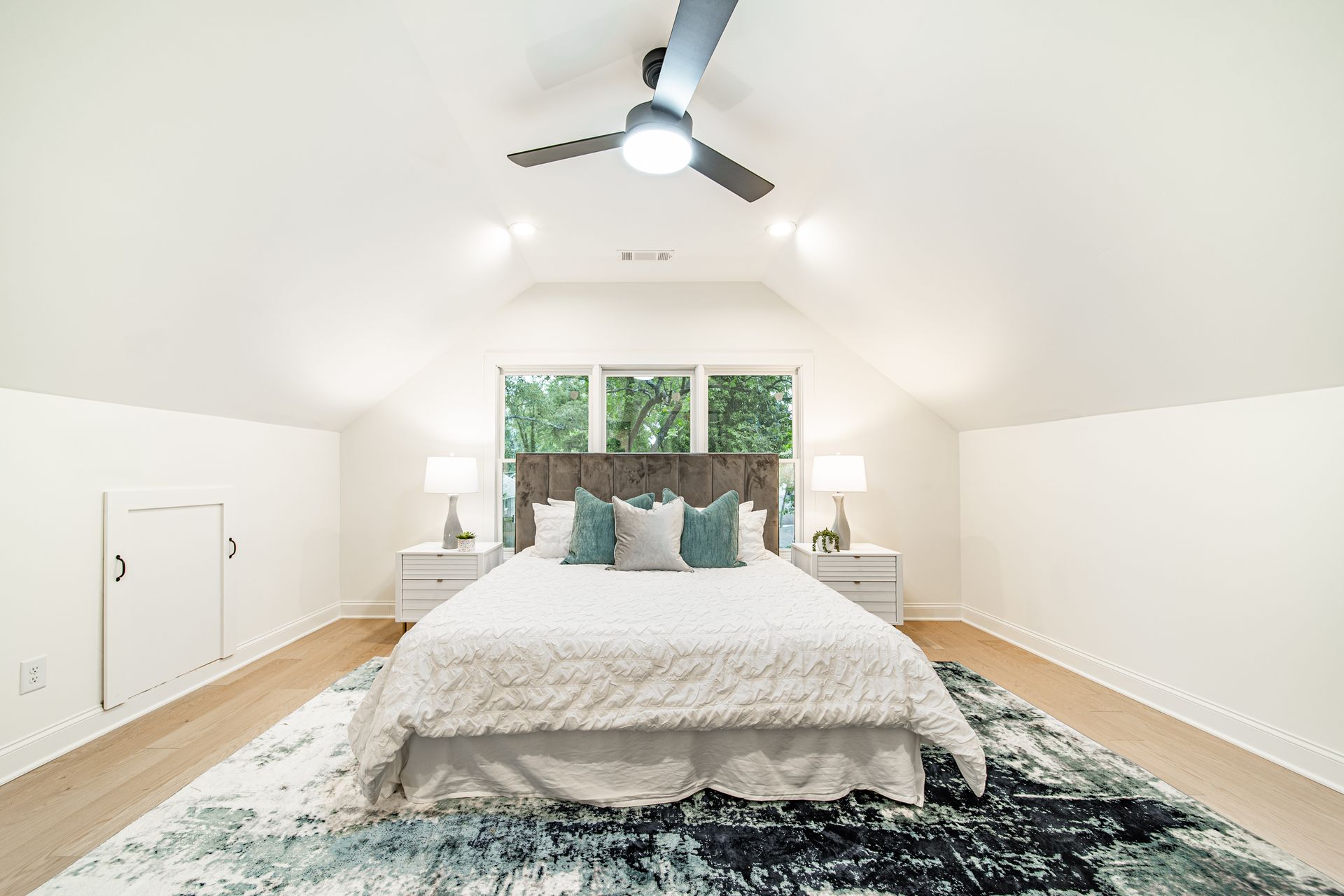 Bedroom with a bed, nightstands, and a large rug. Windows in the background. Ceiling fan.