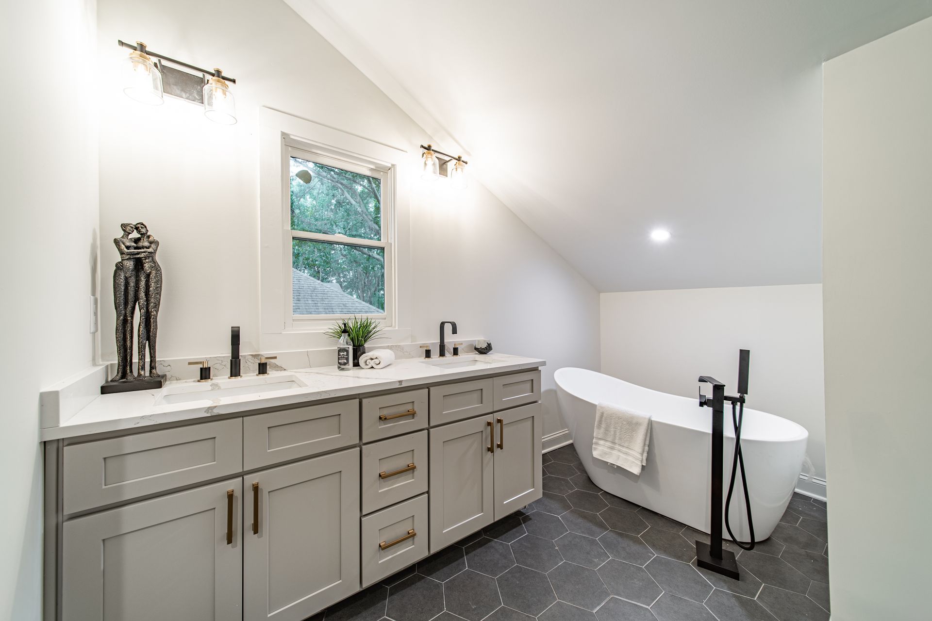 Bathroom with a gray vanity, black fixtures, freestanding tub, and window, set in a sloped ceiling room.
