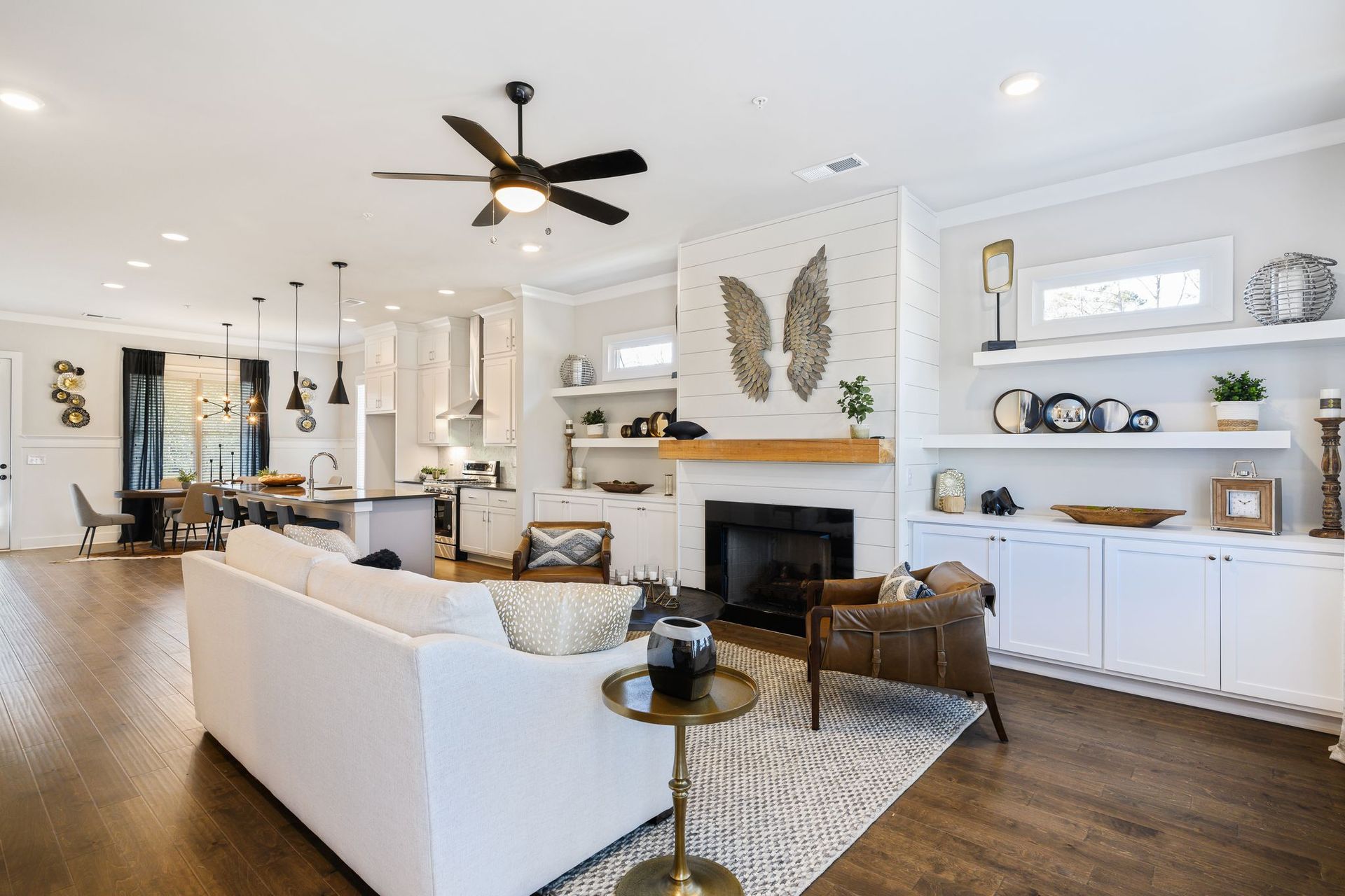 Bright open-concept living space with white walls, dark wood floors, and a fireplace. A white sofa and brown armchair are in the center.