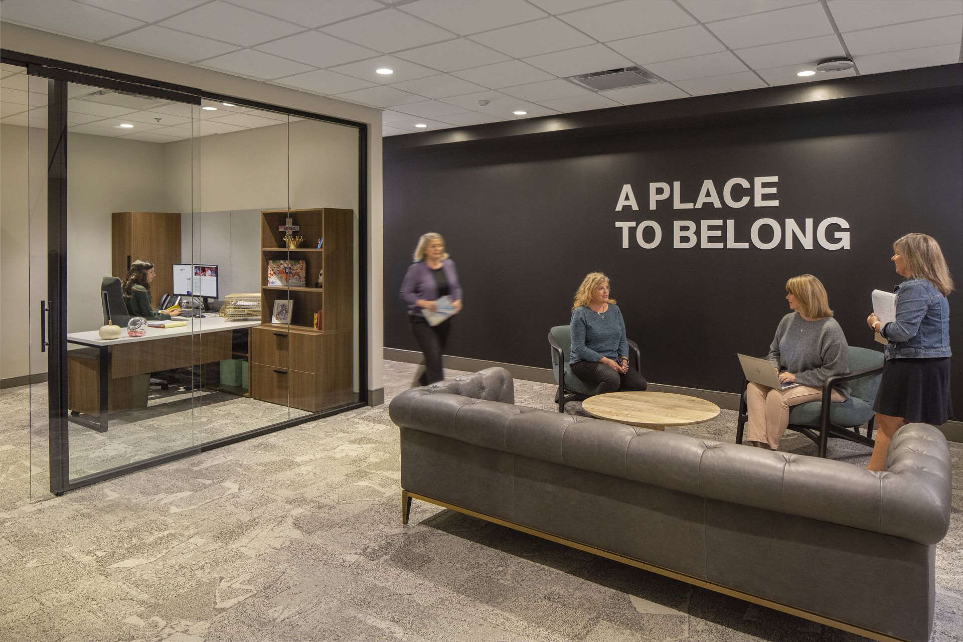 Office interior with people near couch, glass-walled office visible; black wall with text 