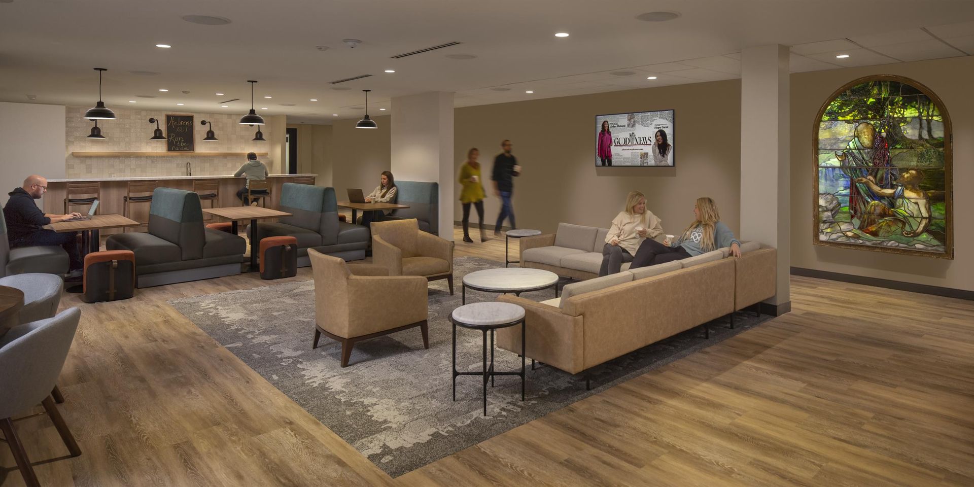 Lounge area with seating, tables, a TV, and a stained glass window. People are present.