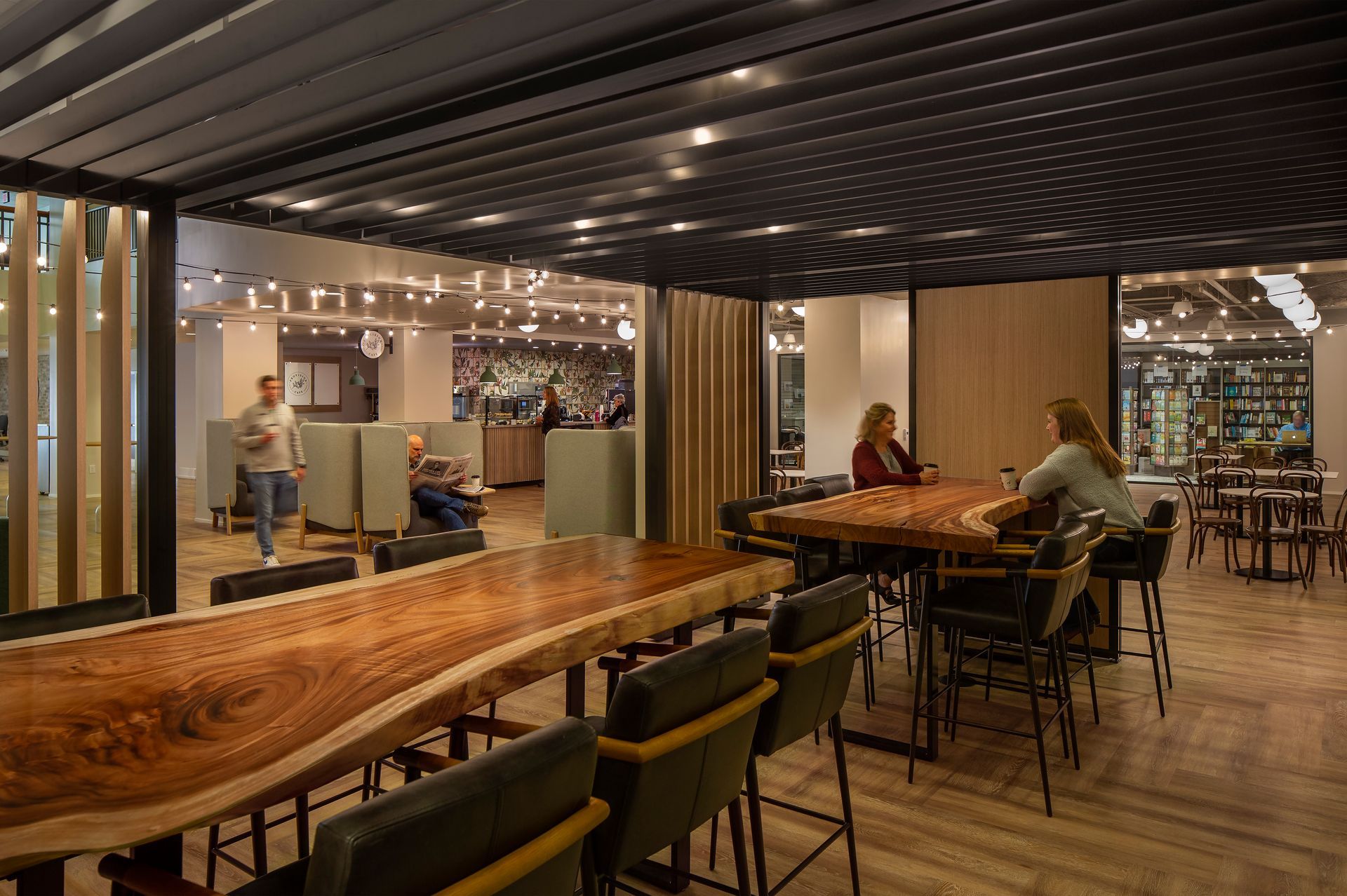 Interior of a modern office space with wooden tables and chairs. People are working and conversing in a well-lit area.
