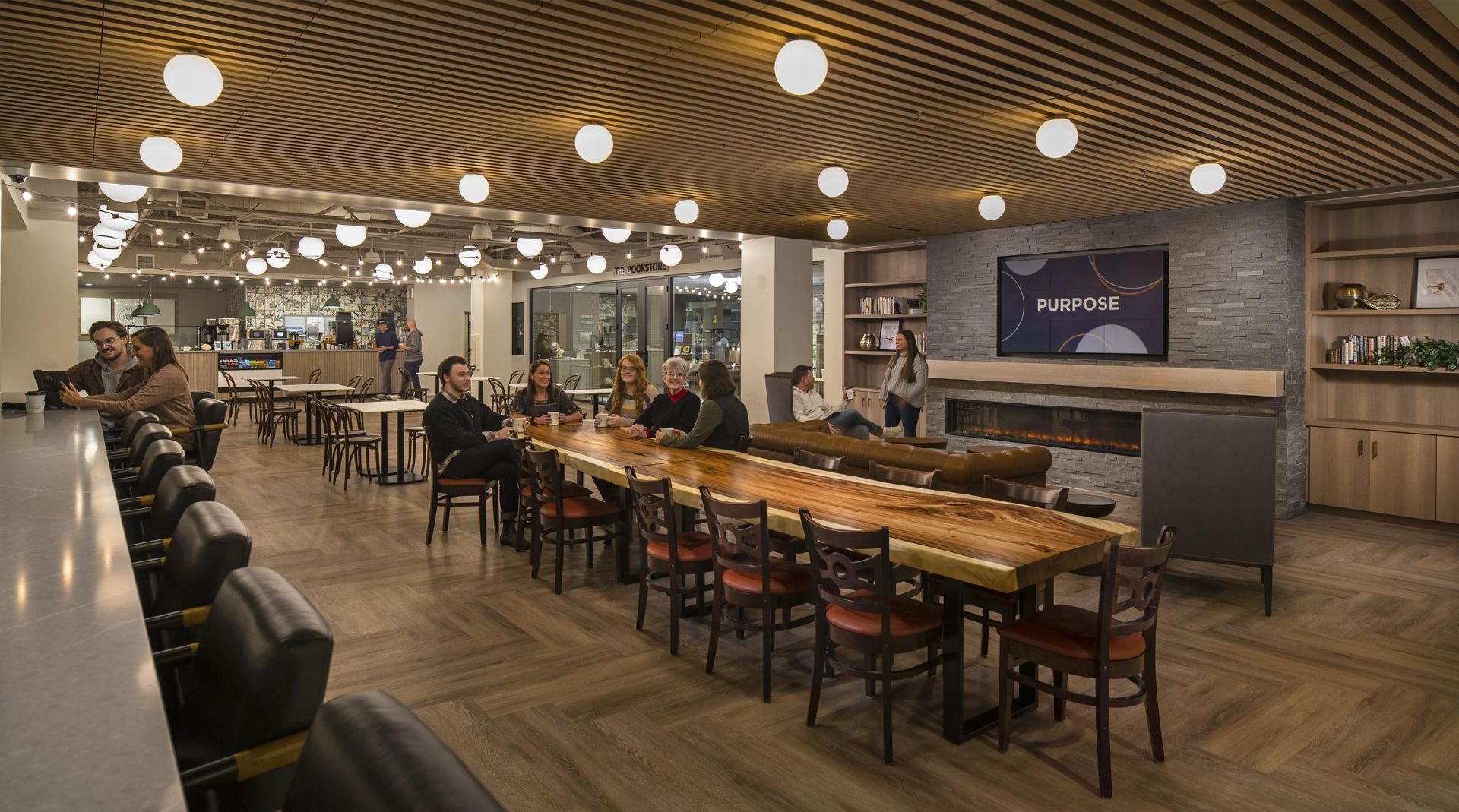 Interior of a modern lounge with a long wooden table, bar seating, and a fireplace. People sit and gather.