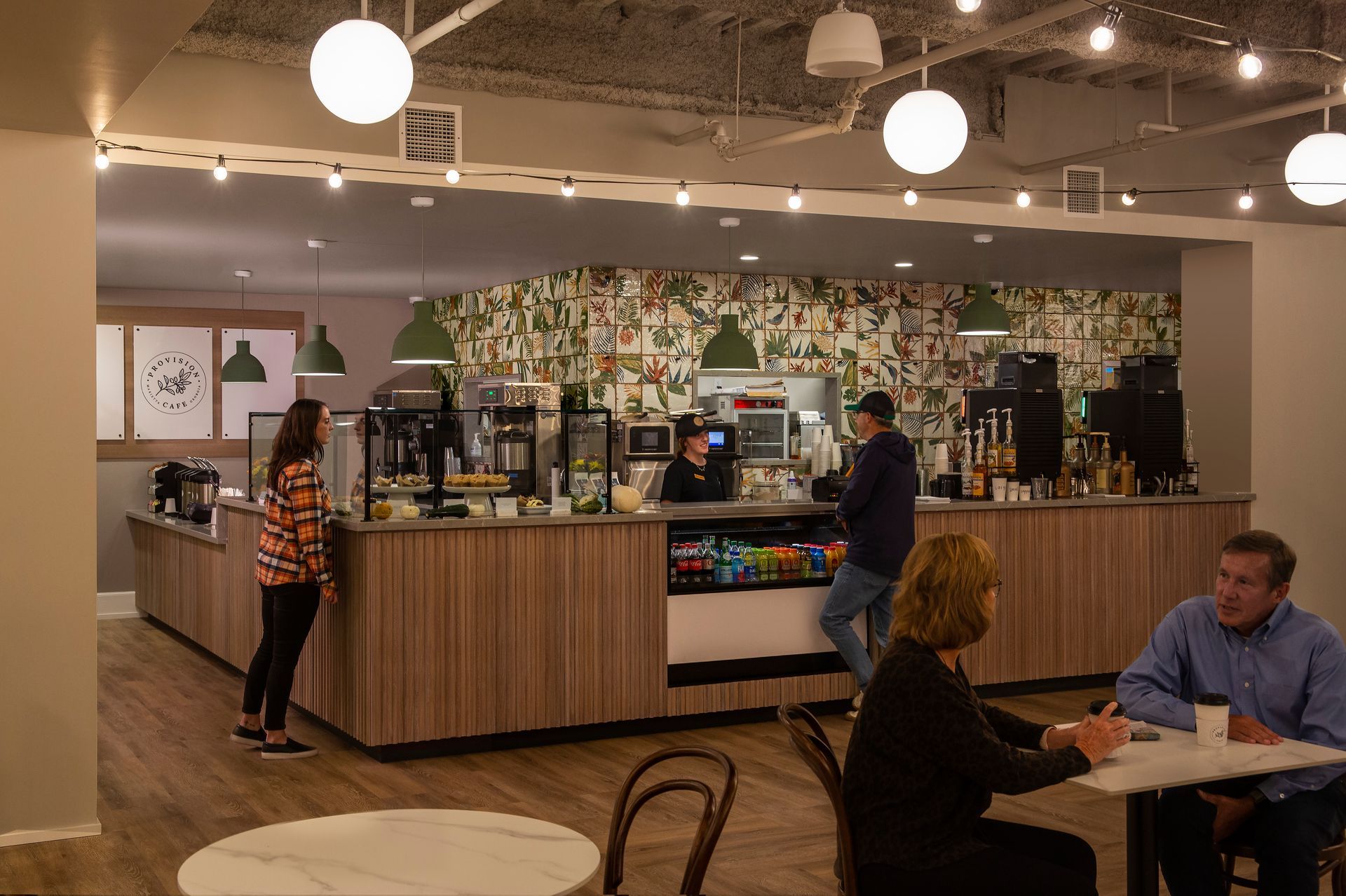 Coffee shop interior: customers near counter; staff serving drinks. Bright lights, wood paneling, decorative wall.