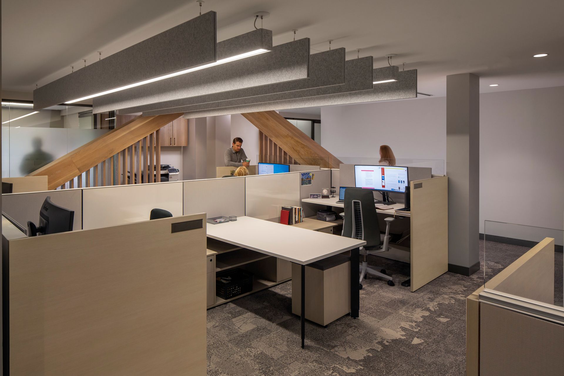 Office interior with cubicles, desks, and two people near a wooden staircase.