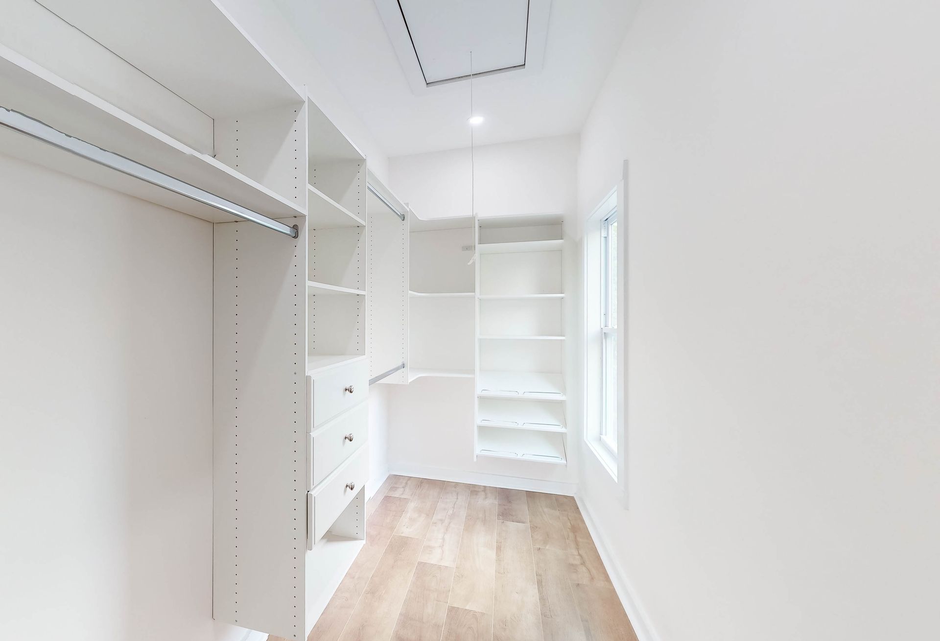 Empty walk-in closet with white shelves, drawers, and wooden floor. A small window lets in light.