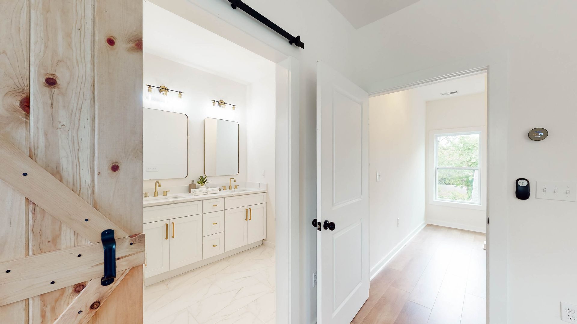 Barn door leading to bathroom, white cabinets, gold hardware. Hallway to bright window with thermostat.