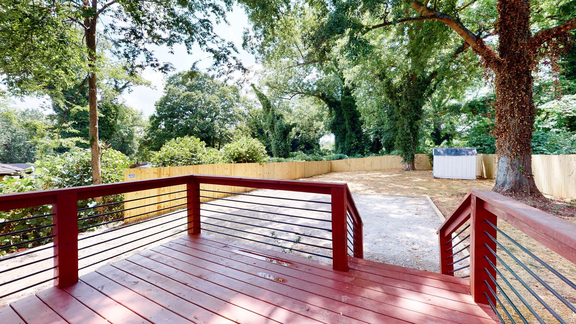 Wooden deck with red railings overlooking a gravel yard, backed by a wooden fence and lush trees.