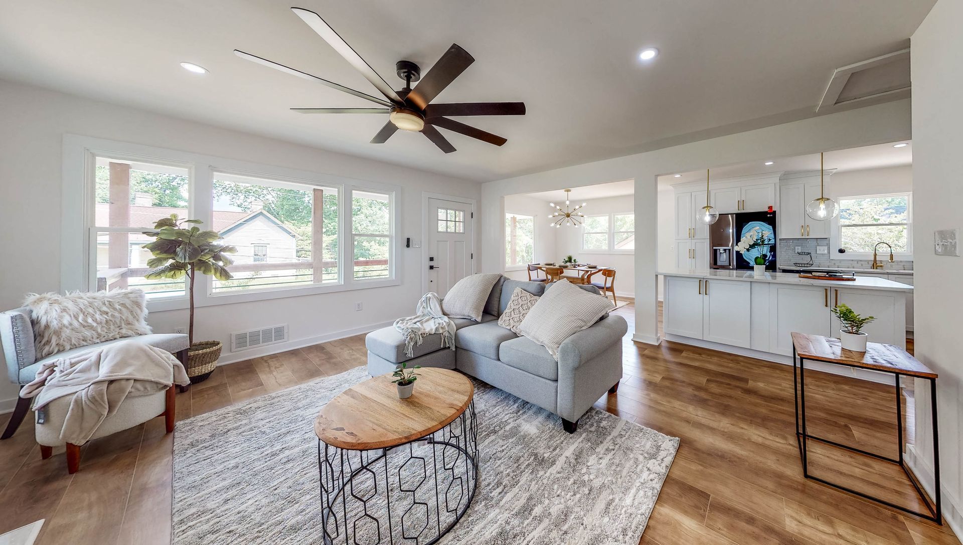 Bright living room with gray couch, rug, and wooden floors. Open to a kitchen.