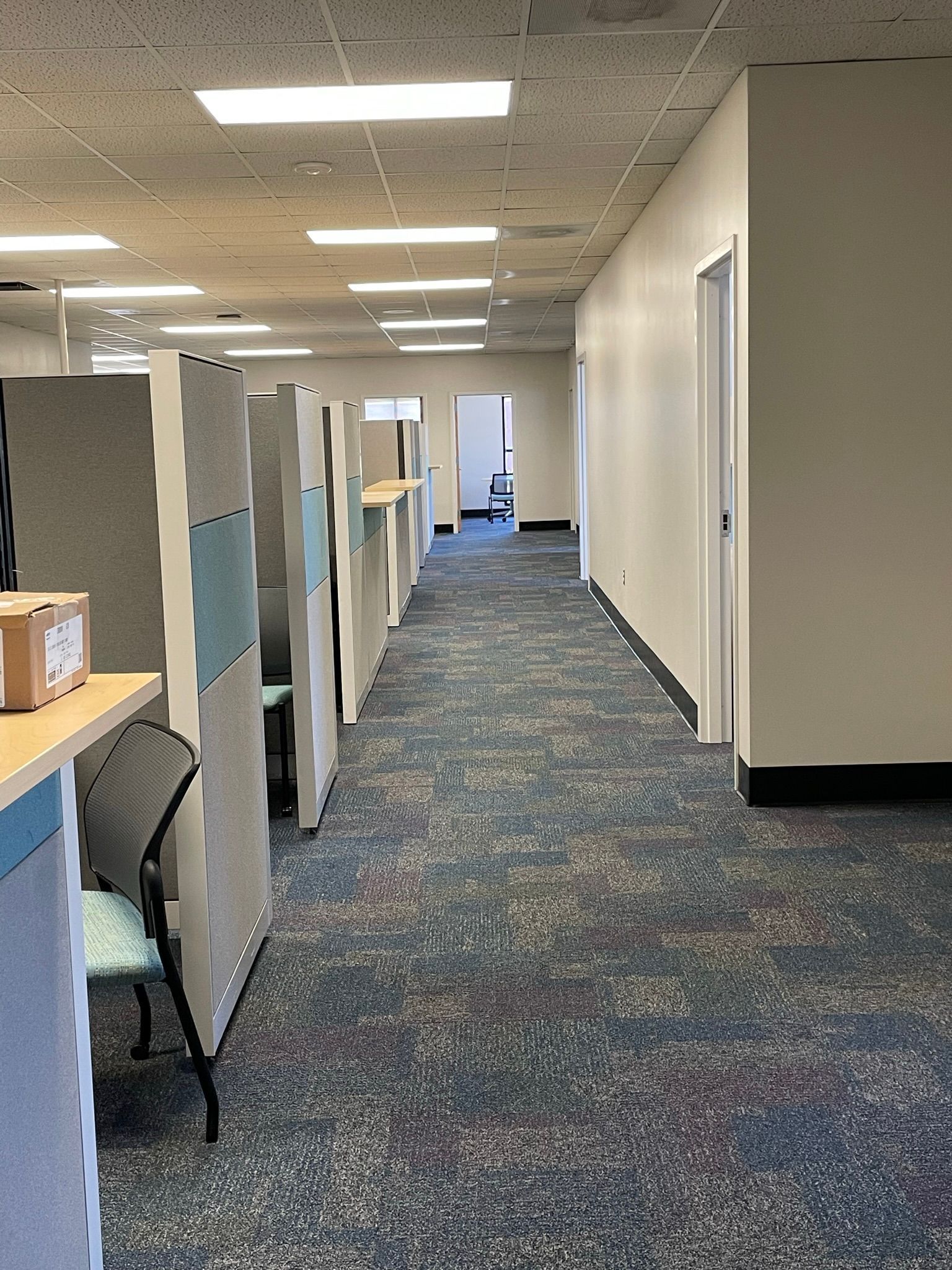 Office hallway with cubicles on the left and a wall with doors on the right. Blue and gray carpet.
