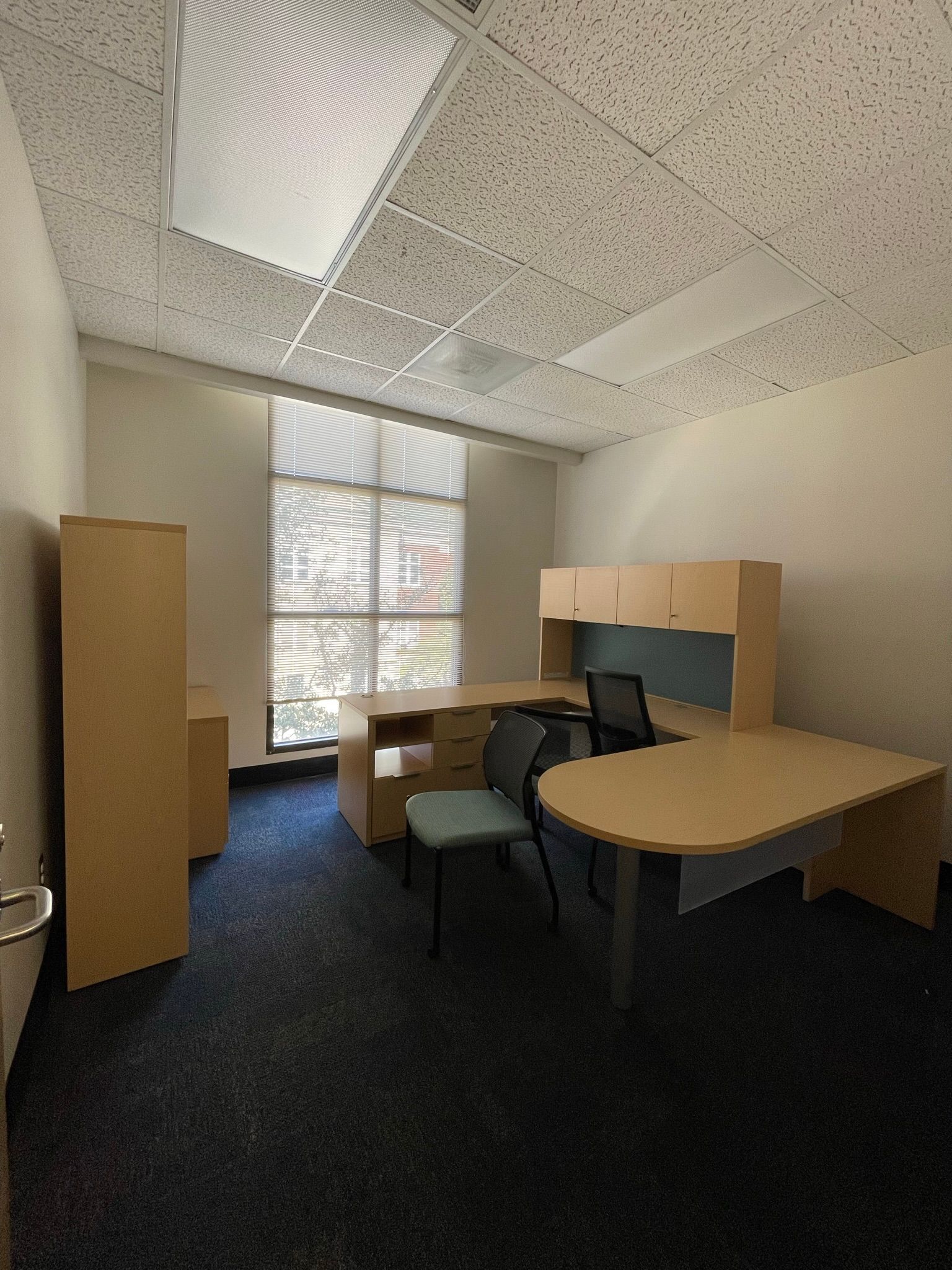 Office with beige furniture, blue carpet, and a window. Cabinets, desks, and chairs are present. Ceiling tiles visible.