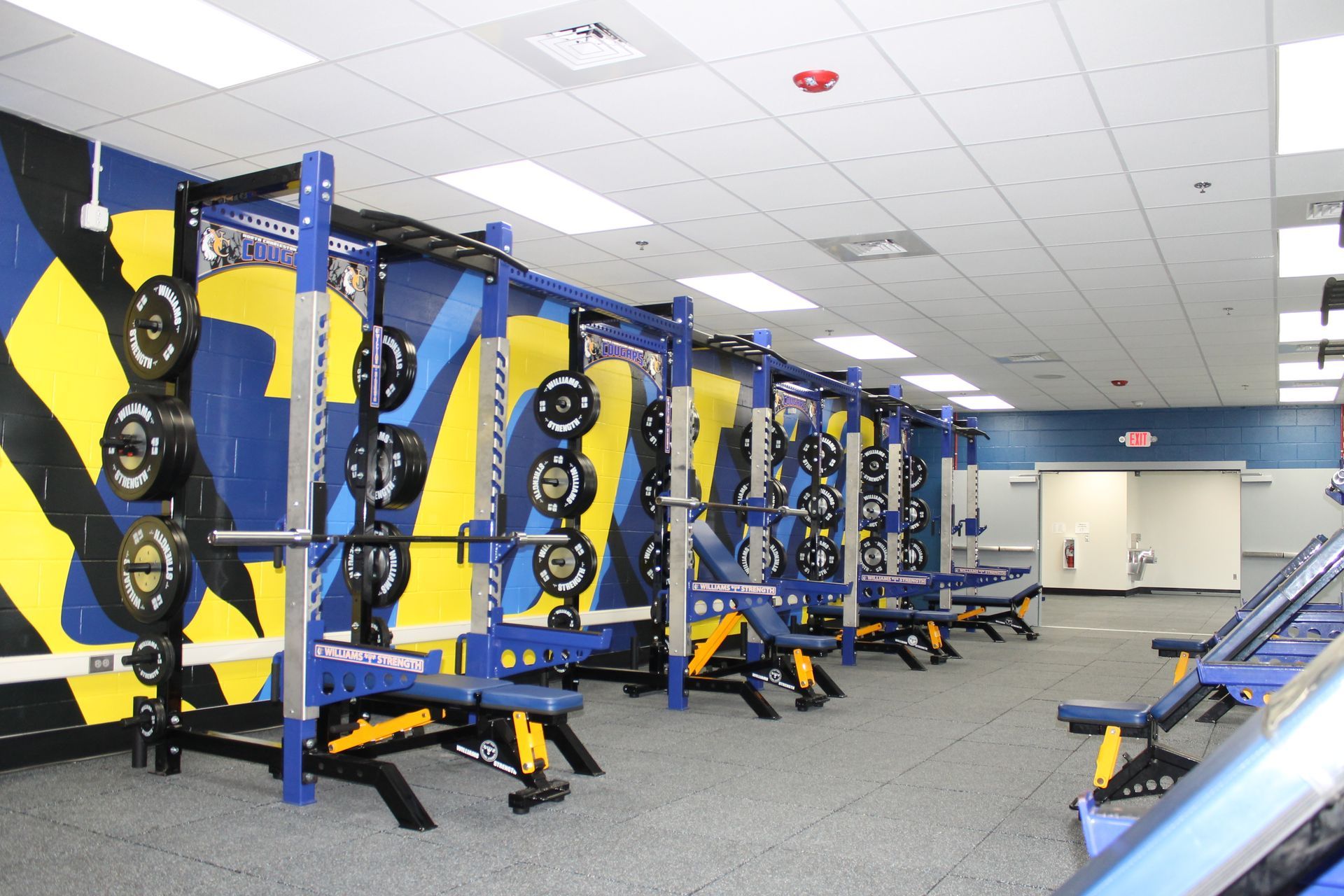 Weight room with blue and yellow weight racks, benches, and weights.
