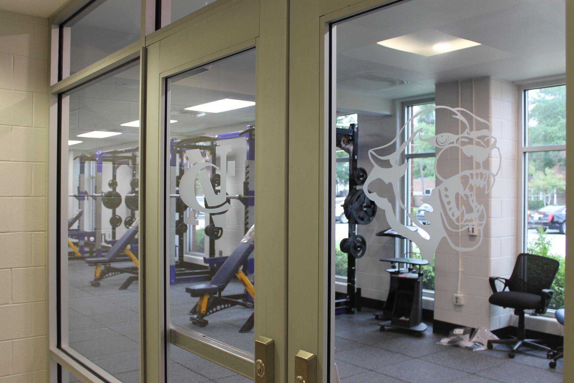 Weight room viewed through glass doors with etched design; gym equipment visible.