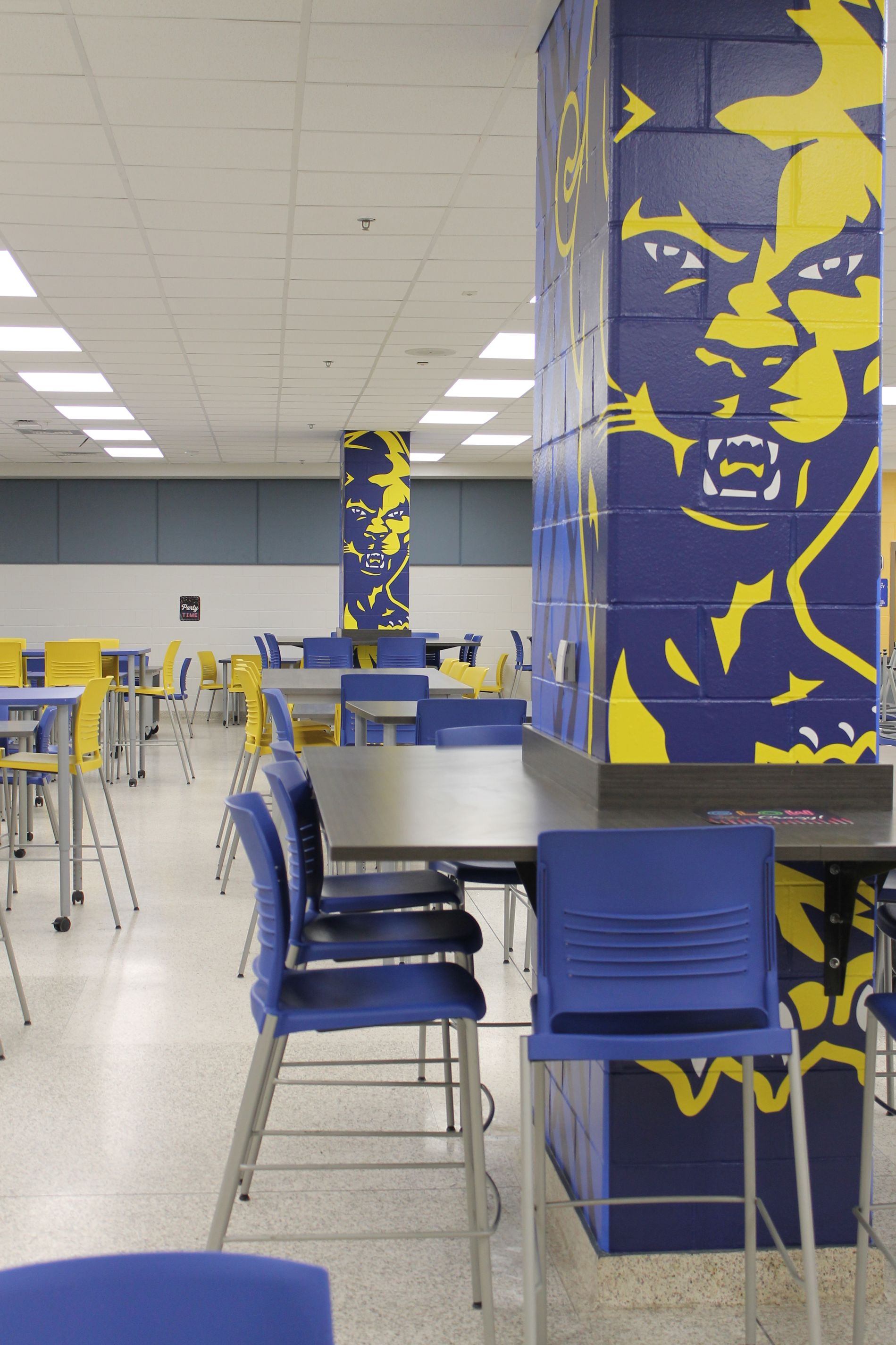 School cafeteria with blue and yellow chairs and columns featuring a lion graphic.