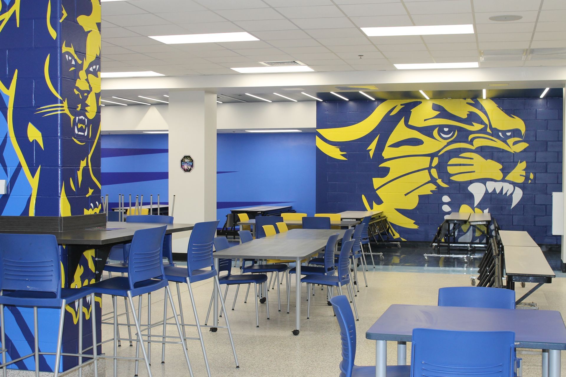 School cafeteria with blue and yellow cougar mascot mural and tables with blue chairs.