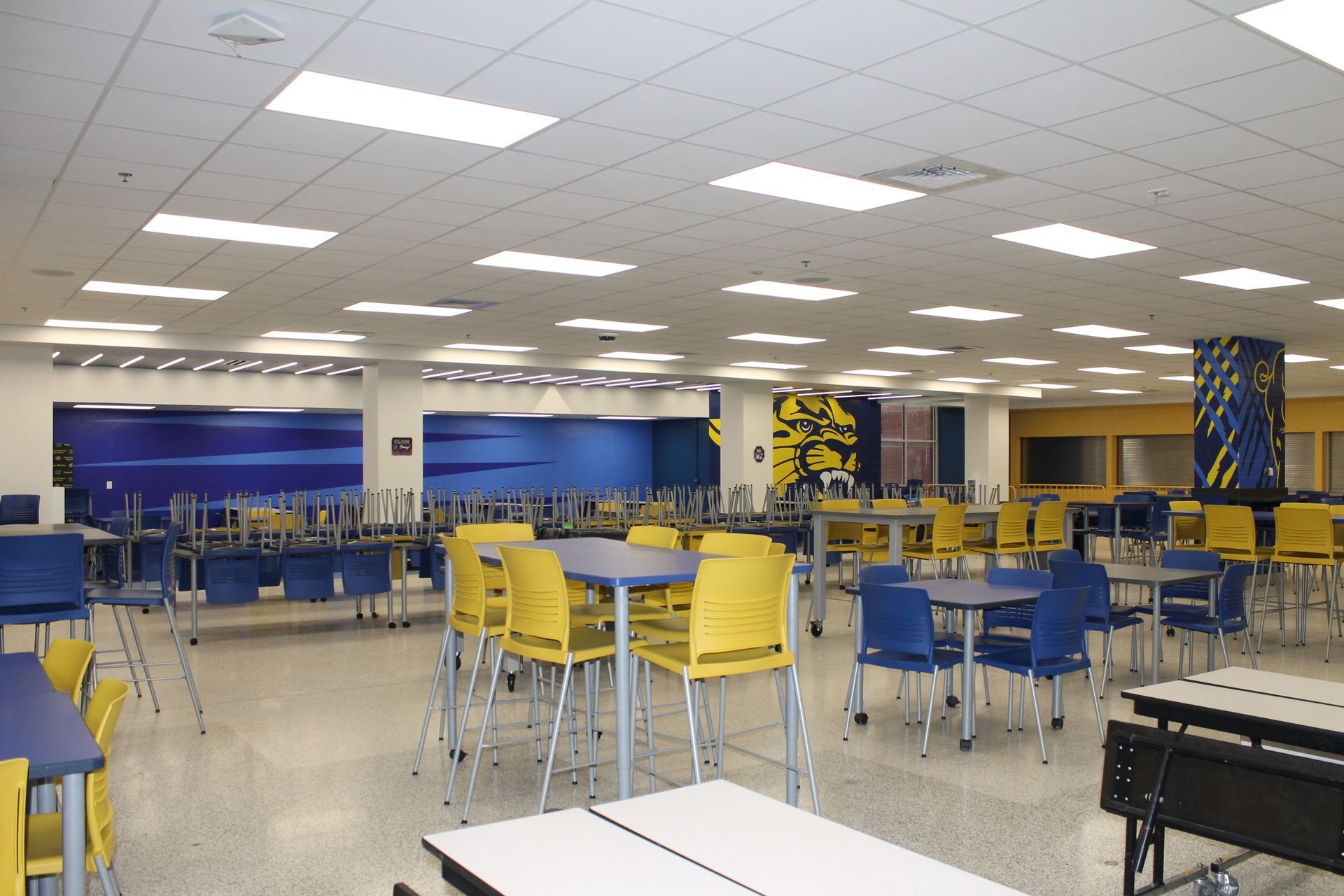 Empty school cafeteria with blue and yellow chairs and tables, blue wall, overhead fluorescent lights.