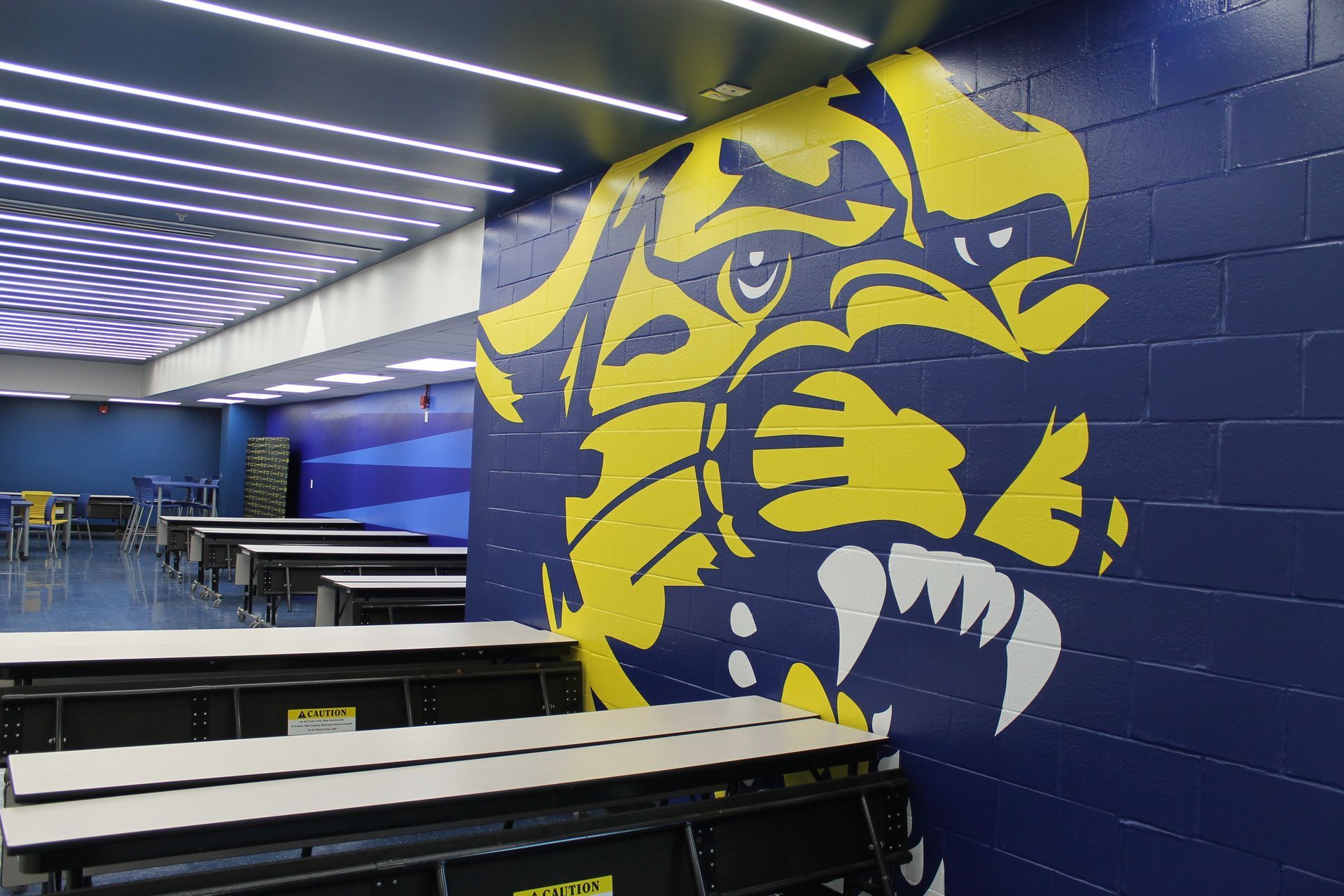 A yellow and blue tiger mascot painted on a blue brick wall in a school cafeteria.