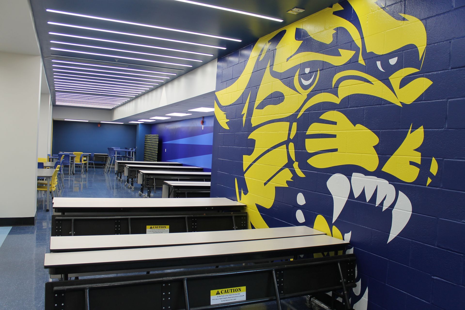School cafeteria with blue and yellow tiger mascot mural. Rows of tables and chairs.