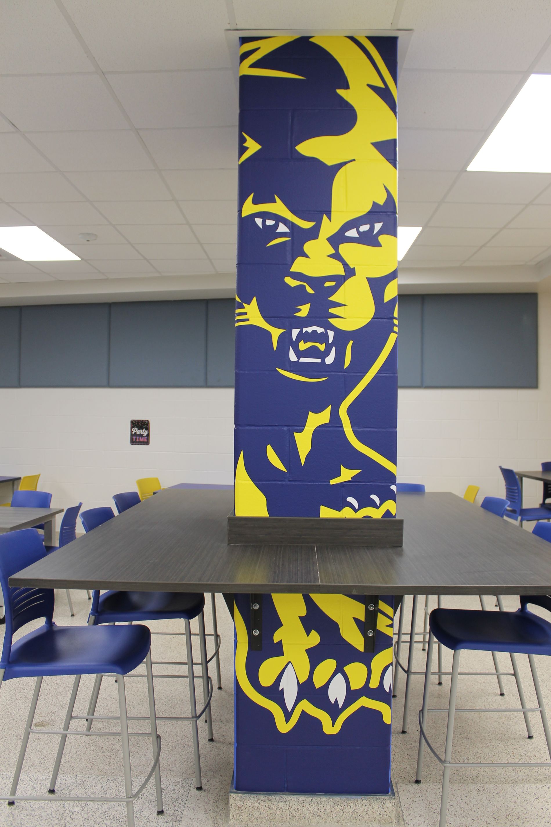 A blue and yellow panther mascot mural on a support column in a school cafeteria, surrounded by tables and chairs.