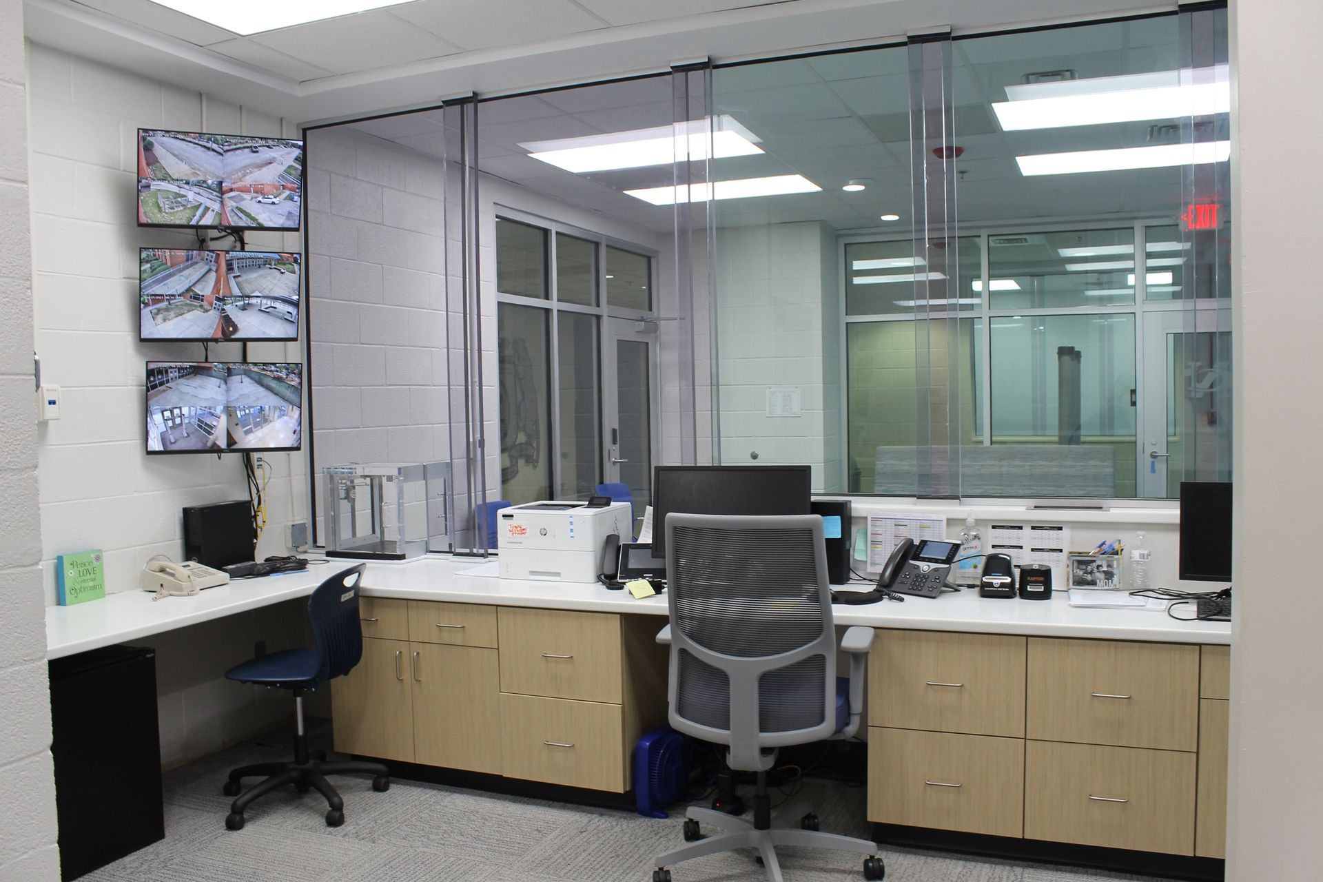 Security control room with monitors displaying camera feeds, desks, and a clear glass partition.