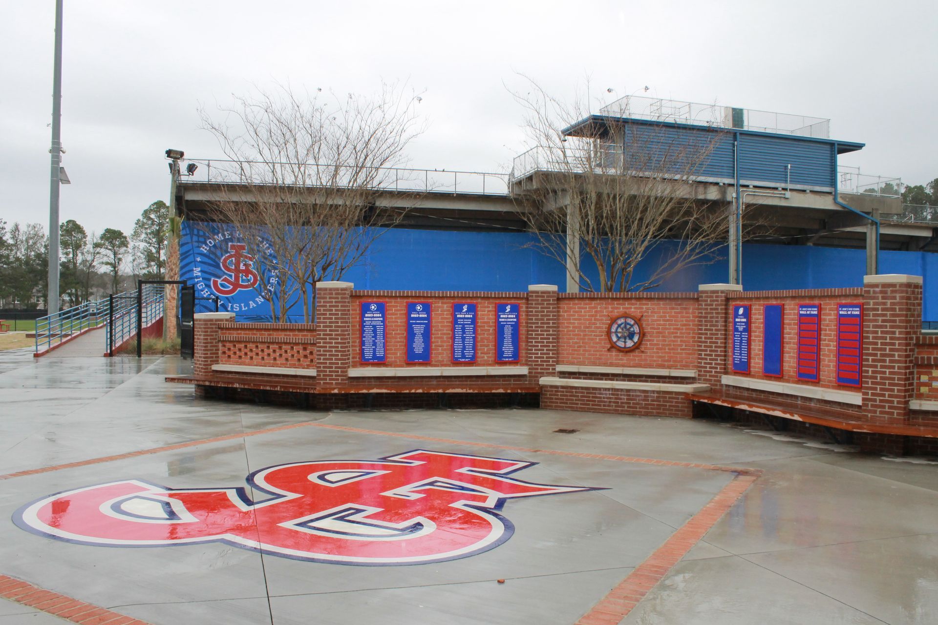 Brick memorial wall with plaques, blue backdrop, logo in foreground, cloudy day.