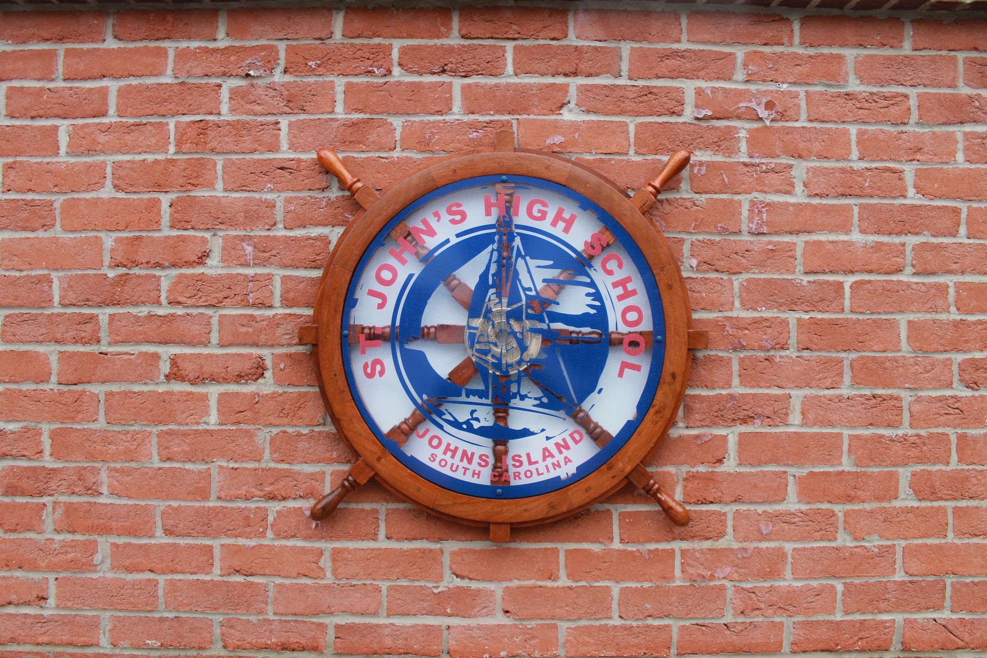 St. John's High School sign, nautical wheel design, mounted on a red brick wall.