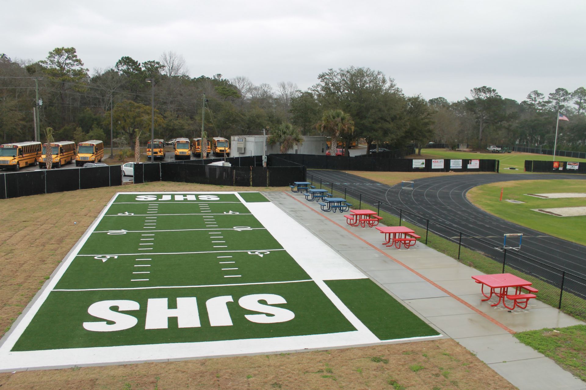 Football field with green turf, track, school buses, and picnic tables; overcast day.