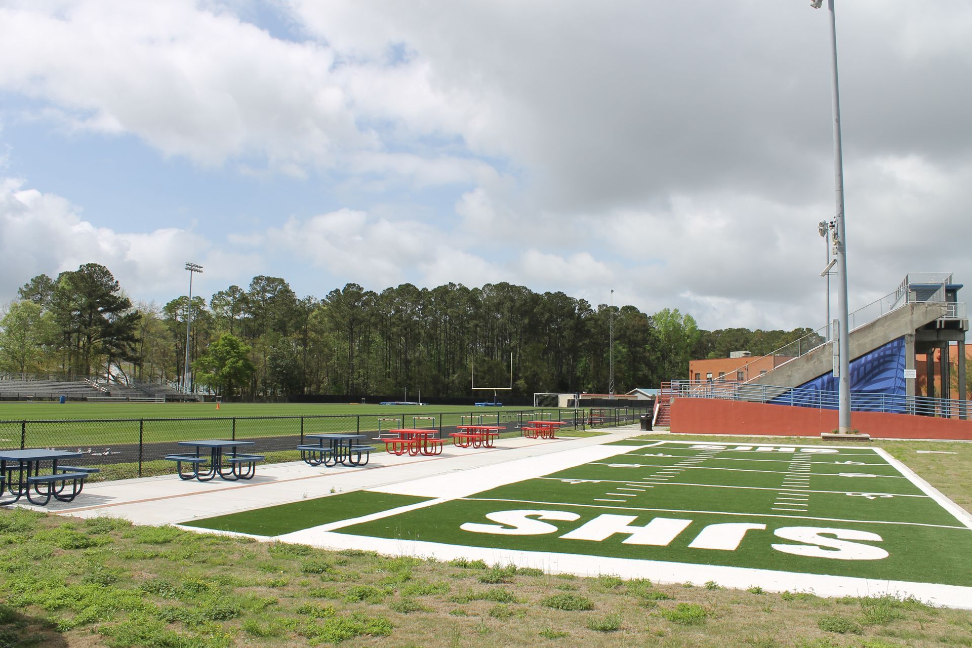 Football field with bleachers, picnic tables, and trees under a cloudy sky. 