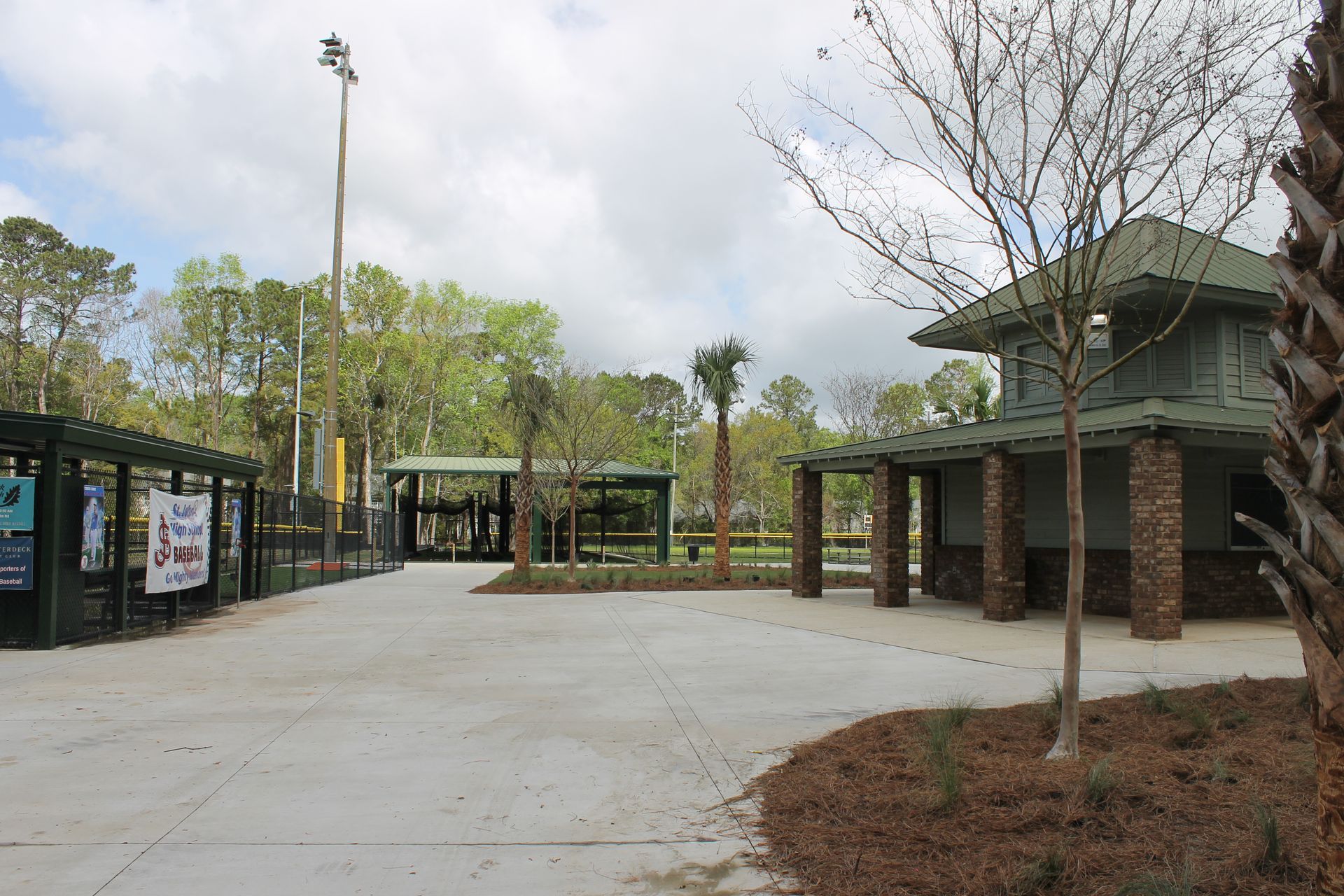 Entrance to a baseball field complex, featuring a concrete path, a covered structure, and a baseball field in the background under a cloudy sky.